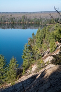 A view from the top of a sand dune. The calm blue water of the river is in the distance, with pine trees reflecting from the far shore. 